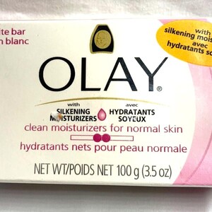 Puede incluir: Una pastilla de jabón Olay blanca con detalles rosas. El envase presenta el logotipo de Olay, texto que indica que es una pastilla blanca con hidratantes sedosos para piel normal. El peso neto es de 100 g.