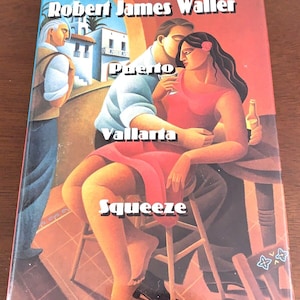 May include: A book cover with the title "Puerto Vallarta Squeeze" by Robert James Waller. The cover art depicts a couple embracing, with a man holding a woman who is holding a drink. The background features a building and another man.