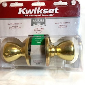 Può includere: Un nuovo set di pomelli per porte Kwikset in una confezione di plastica trasparente. I pomelli sono di colore oro e hanno una forma rotonda. La confezione include il testo "The Beauty of Strength" e "Hall & Closet".