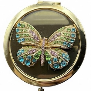 May include: A gold-toned, round compact mirror with a colorful jeweled butterfly design. The butterfly features blue, green, and purple gemstones. The mirror has a black background and a hinged opening.