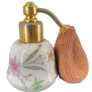 May include: A vintage perfume bottle with a white porcelain body decorated with pink and gold floral designs. The bottle has a gold-colored metal top and a matching sprayer. A peach-colored mesh atomizer is attached to the sprayer.