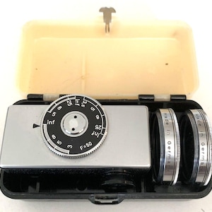 May include: A vintage camera lens kit in a black and beige case. The kit includes a silver camera body with a black dial and two lenses. The lenses are marked "Made in Germany". The dial has numbers and the text "F=50".