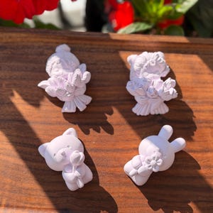 May include: Four small, matte white decorative figurines. Two figurines depict characters holding large bouquets of flowers, one depicts a cat holding a single flower, and one depicts a rabbit holding a flower. The figures have smooth, solid surfaces.