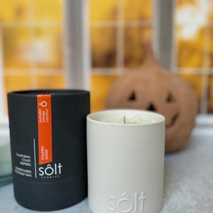 Può includere: Una candela bianca con la parola "sölt" in rilievo sul lato. La candela è in una scatola nera con il testo "sölt candles" e "pumpkin sunset" sul lato. La candela è su una superficie grigia.