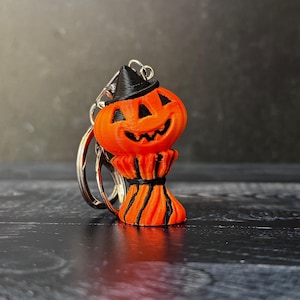 May include: Orange pumpkin keychain with a black witch hat and a smiling face. The pumpkin has a decorative base with black stripes. The keychain is attached to a silver metal ring, perfect for Halloween.