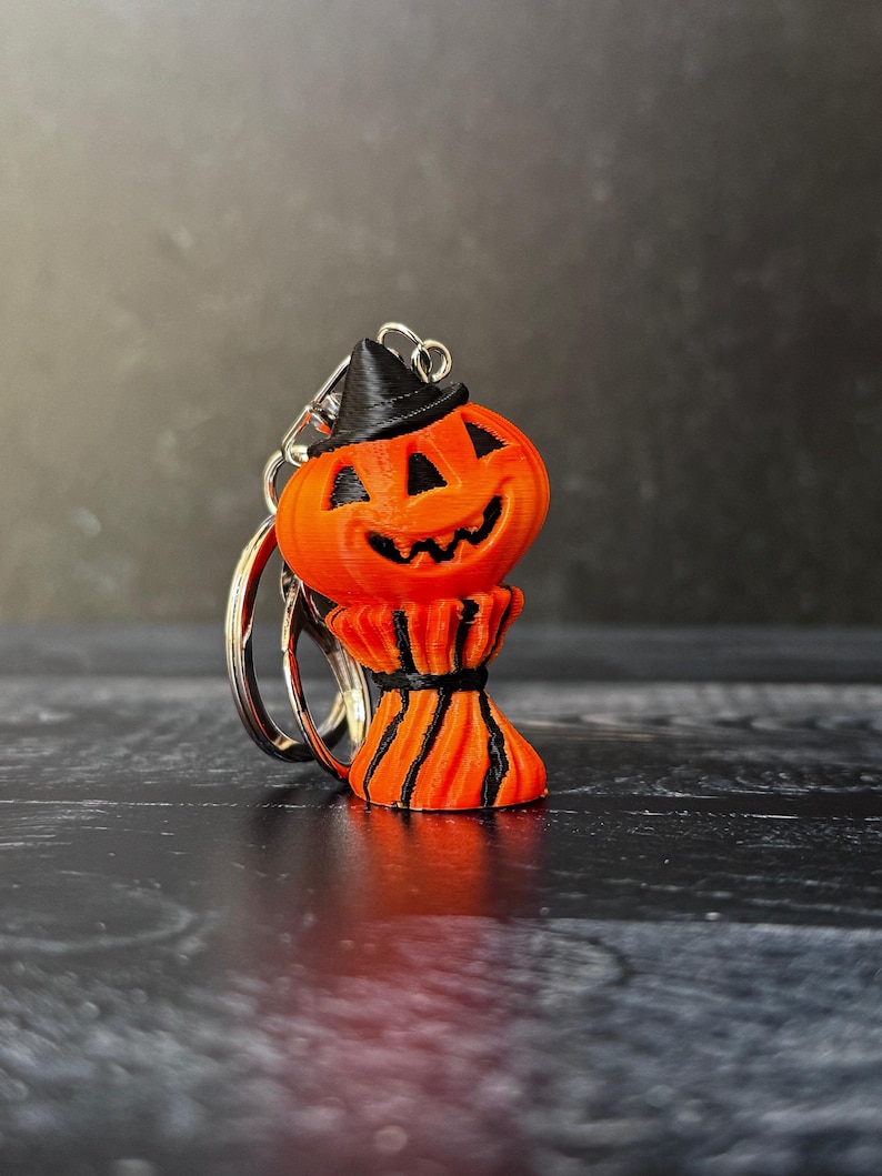 May include: Orange pumpkin keychain with a black witch hat and a smiling face. The pumpkin has a decorative base with black stripes. The keychain is attached to a silver metal ring, perfect for Halloween.