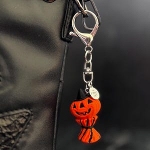 May include: A Halloween-themed pumpkin keychain with a black witch hat and a smiling face. The orange pumpkin charm is attached to a silver-colored clasp and chain, hanging from a black bag.