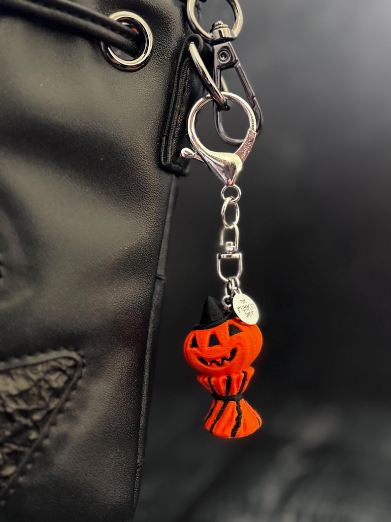 May include: A Halloween-themed pumpkin keychain with a black witch hat and a smiling face. The orange pumpkin charm is attached to a silver-colored clasp and chain, hanging from a black bag.