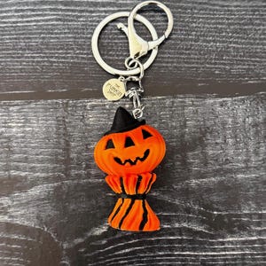 May include: A Halloween-themed keychain featuring an orange pumpkin with a black witch hat and a smiling face. The pumpkin is attached to a silver keyring with a lobster clasp. The keychain has a small circular tag that says "The Funkin Shop".