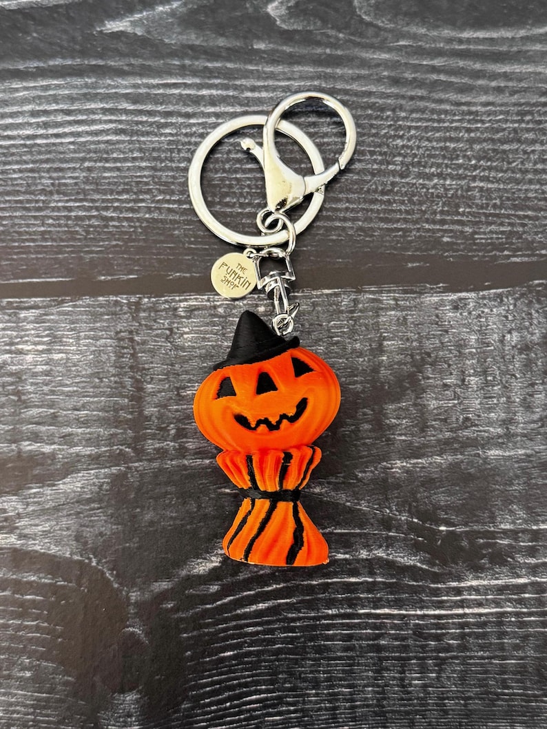 May include: A Halloween-themed keychain featuring an orange pumpkin with a black witch hat and a smiling face. The pumpkin is attached to a silver keyring with a lobster clasp. The keychain has a small circular tag that says "The Funkin Shop".
