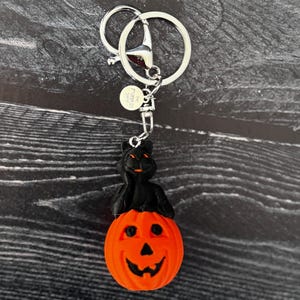 May include: A Halloween-themed keyring featuring a black cat perched atop an orange pumpkin with a carved face. The keyring has a silver-coloured ring and clasp for attaching to keys or bags.