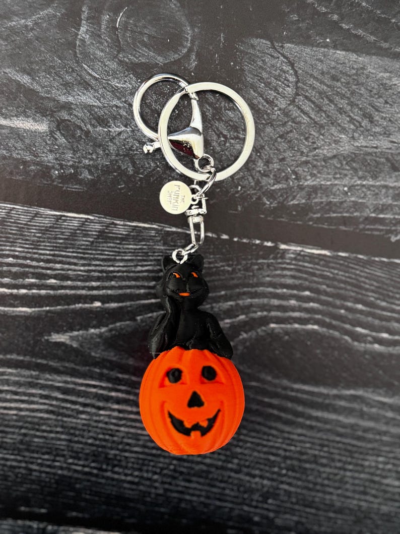 May include: A Halloween-themed keyring featuring a black cat perched atop an orange pumpkin with a carved face. The keyring has a silver-coloured ring and clasp for attaching to keys or bags.