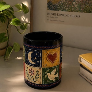 Puede incluir: Taza de cerámica negra con un diseño de arte popular colorido. El diseño incluye una luna creciente y estrellas, un corazón rojo, una flor y una paloma. La taza está decorada con un borde ondulado azul.
