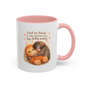 Könnte beinhalten: Weißer Keramikbecher mit rosa Innenraum und Henkel. Der Becher zeigt eine Illustration von zwei sich umarmenden Affen mit dem Text "I feel so loved, it's like my heart got a big, fluffy punch."