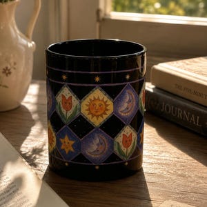 May include: Black ceramic mug with a repeating pattern of celestial and floral designs. The pattern includes a smiling sun, crescent moons, stars, and tulips in a diamond grid. The mug sits on a wooden surface near a window.