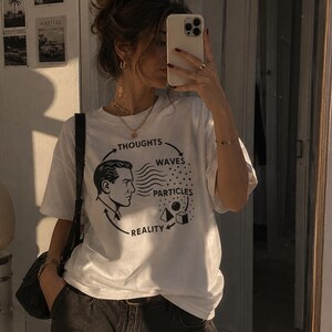 Könnte beinhalten: Weißes T-Shirt mit einem grafischen Design, das das Profil eines Mannes, Wellen, Partikel und die Worte "Gedanken, Wellen, Partikel, Realität" zeigt. Das Shirt wird mit schwarzen Jeans und einer schwarzen Umhängetasche kombiniert. Das Bild ist in einem gut beleuchteten Raum aufgenommen.