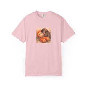 Könnte beinhalten: Hellrosa T-Shirt mit einem Motiv von zwei sich umarmenden Affen. Einer ist braun, der andere orange. Das Motiv ist mittig platziert, mit einem kleinen Etikett am Halsausschnitt.
