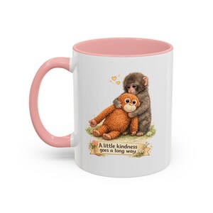 Könnte beinhalten: Weiße Keramik-Tasse mit rosa Innenraum und Henkel. Die Tasse zeigt eine Illustration eines Affen, der einen Stofforang-Utan umarmt. Der Text "A little kindness goes a long way" ist auf einem Banner unter der Illustration aufgedruckt.