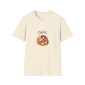 Könnte beinhalten: Cremefarbenes T-Shirt mit einer Grafik von zwei sich umarmenden Zeichentrickaffen. Der Text "I feel so loved, it's like my heart got a big, fluffy punch" steht über den Affen. Das Shirt ist aus weichem Material.