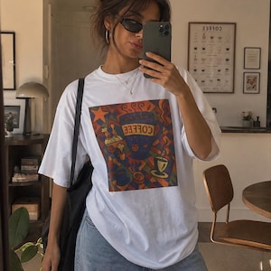 Puede incluir: Camiseta blanca con un gráfico colorido con temática de café. El diseño presenta una taza de café, vapor y la palabra "COFFEE" en una fuente estilizada. La camiseta se combina con vaqueros azules y un bolso tote negro.