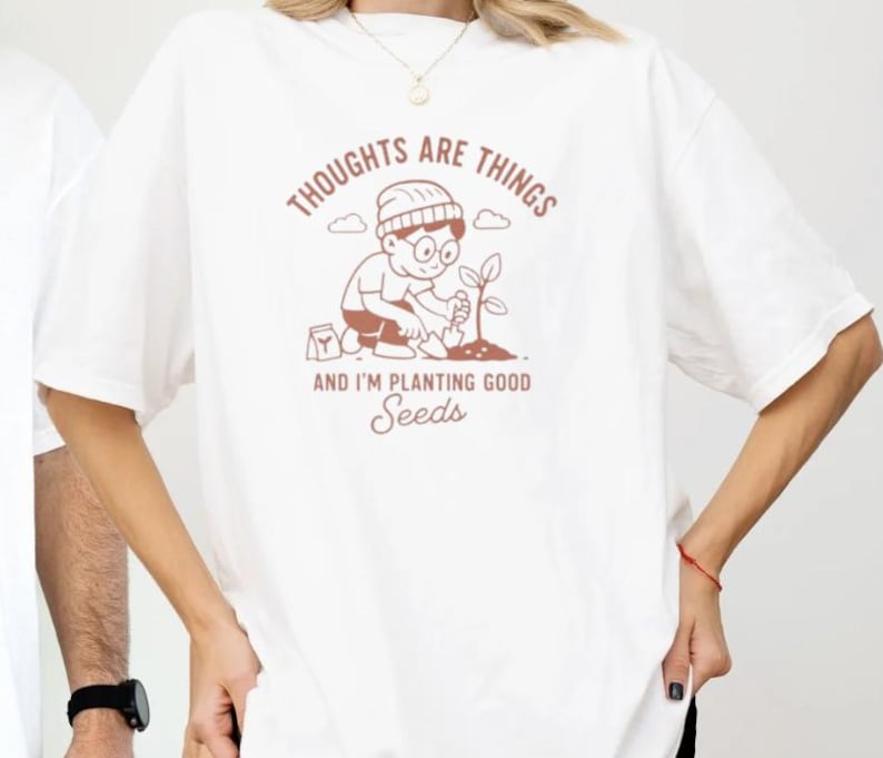 May include: White t-shirt with a brown graphic of a person planting a seedling, with the text "THOUGHTS ARE THINGS" above and "AND I'M PLANTING GOOD SEEDS" below. The shirt is a casual, oversized fit.