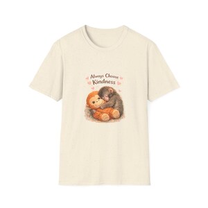 Könnte beinhalten: Cremefarbenes T-Shirt mit einem Aufdruck von zwei sich umarmenden Stofftieren, einem braunen Affen und einem grauen Koala. Der Text "Always Choose Kindness" und rosa Herzen sind darüber. Das Shirt ist aus weichem Material.