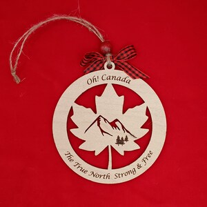Pode incluir: Ornamento de madeira em forma de folha de bordo com uma cena de montanha no interior. O ornamento &eacute; pintado em tons de madeira natural e tem o texto "Oh! Canada The True North Strong & Free" ao redor da borda.