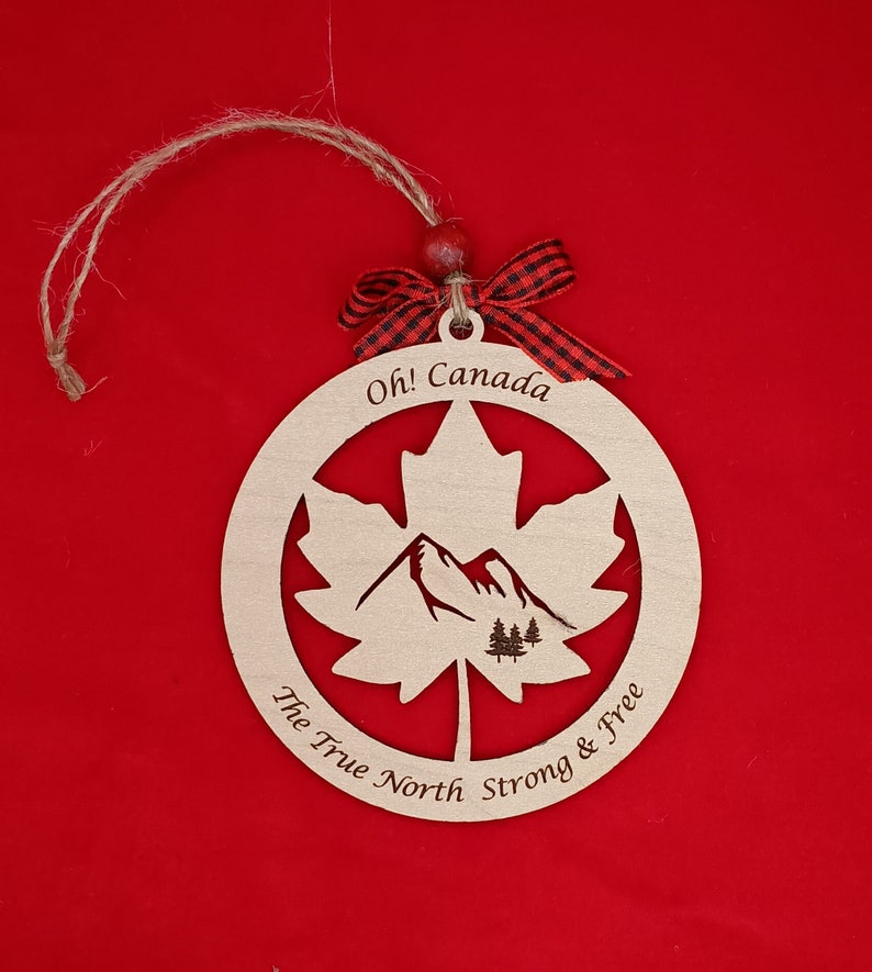 Pode incluir: Ornamento de madeira em forma de folha de bordo com uma cena de montanha no interior. O ornamento &eacute; pintado em tons de madeira natural e tem o texto "Oh! Canada The True North Strong & Free" ao redor da borda.