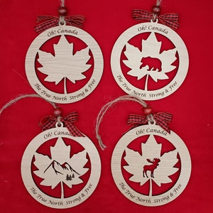 Pode incluir: Quatro ornamentos de madeira em forma de folhas de bordo com diferentes desenhos recortados no centro. Os ornamentos s&atilde;o decorados com uma fita xadrez vermelha e preta e o texto "Oh! Canada The True North Strong & Free".