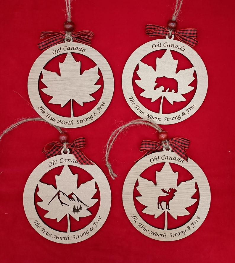 Pode incluir: Quatro ornamentos de madeira em forma de folhas de bordo com diferentes desenhos recortados no centro. Os ornamentos s&atilde;o decorados com uma fita xadrez vermelha e preta e o texto "Oh! Canada The True North Strong & Free".