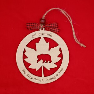 Pode incluir: Ornamento de madeira com uma folha de bordo com um urso dentro, o texto "Oh! Canada The True North Strong & Free" est&aacute; inscrito em torno da folha.