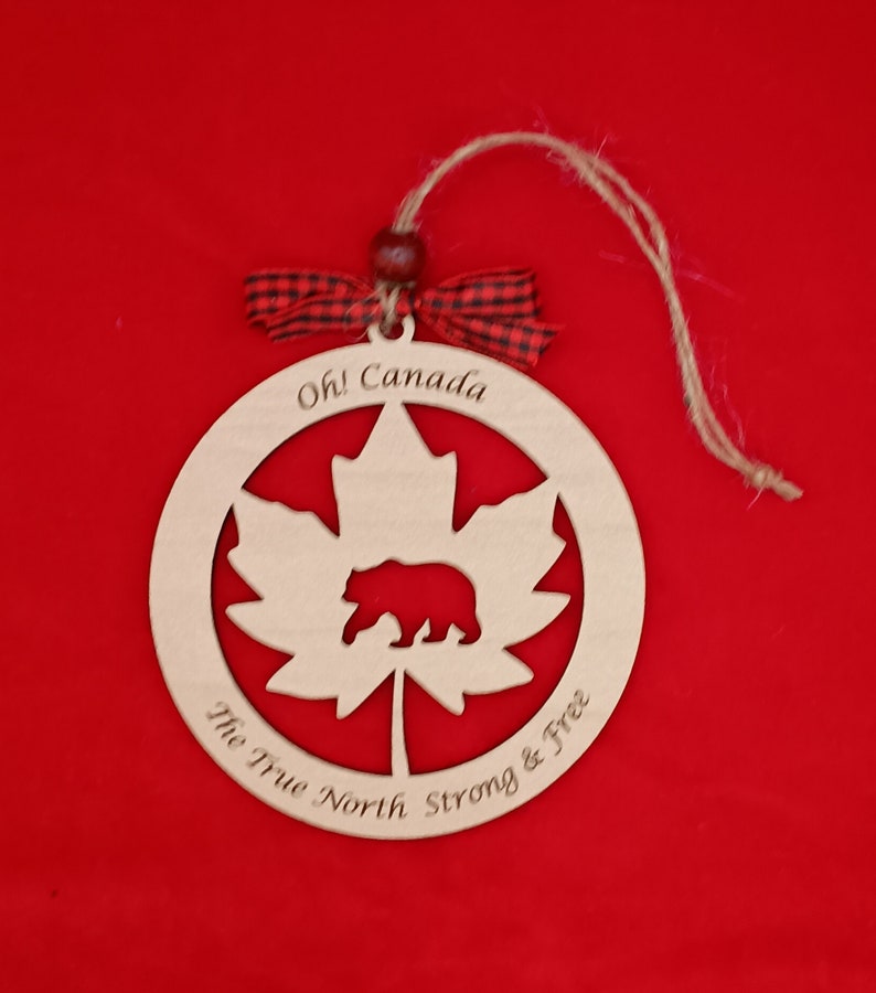 Pode incluir: Ornamento de madeira com uma folha de bordo com um urso dentro, o texto "Oh! Canada The True North Strong & Free" est&aacute; inscrito em torno da folha.