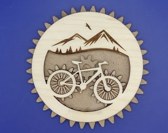 Pode incluir: Uma decoração de parede de madeira com uma cena de montanha com uma silhueta de bicicleta. A bicicleta tem um design de engrenagem e está colocada sobre um fundo castanho.