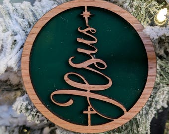 May include: Wooden Christmas ornament with a dark green center and a wooden frame. The word "Jesus" is cut out of the wood in a stylized font, with a star and cross incorporated into the design. The ornament hangs from a jute string.