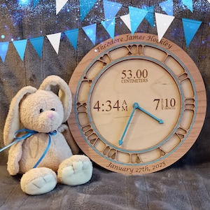 Könnte beinhalten: Eine Holzuhr mit römischen Ziffern, mit dem Namen "Theodore James Hockley", Uhrzeit, Datum und Babymessungen: 53,00 Zentimeter und 7 lbs 10 oz. Ein Plüschhase mit blauem Band sitzt neben der Uhr.