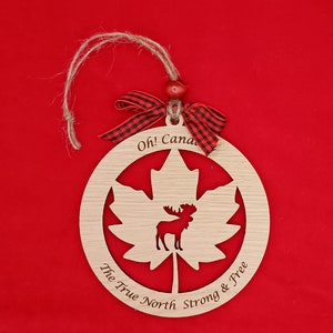 Pode incluir: Ornamento de madeira em forma de folha de bordo com uma silhueta de alce dentro. O ornamento tem o texto "Oh! Canada The True North Strong & Free" ao redor da borda.