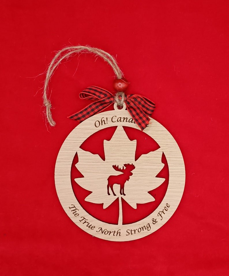 Pode incluir: Ornamento de madeira em forma de folha de bordo com uma silhueta de alce dentro. O ornamento tem o texto "Oh! Canada The True North Strong & Free" ao redor da borda.