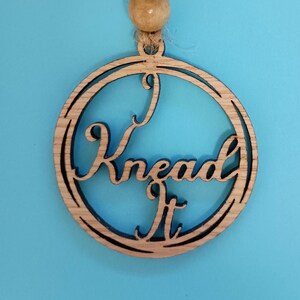 Puede incluir: Un adorno redondo de madera con las palabras "I Knead It" recortadas en una fuente cursiva. El adorno está unido a una cuerda marrón y tiene una cuenta de madera en la parte superior.