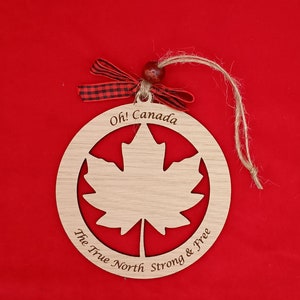 Pode incluir: Ornamento de madeira em forma de c&iacute;rculo com um recorte de folha de bordo. O ornamento &eacute; pintado de marrom e tem o texto "Oh! Canada The True North Strong & Free" inscrito ao redor da borda.