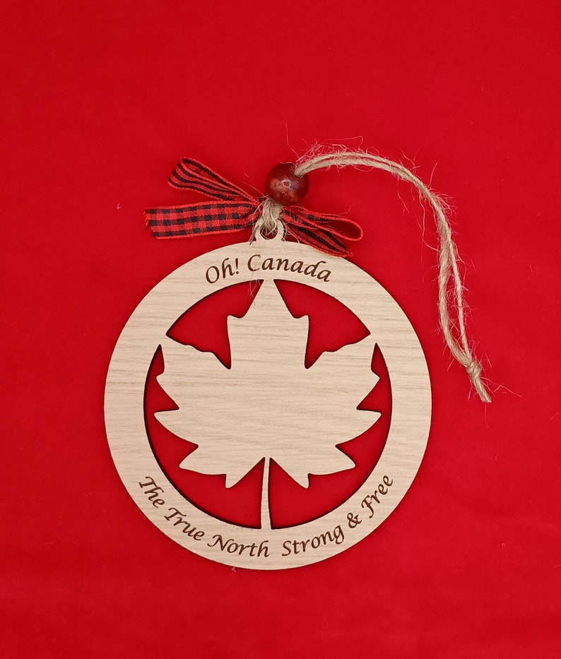 Pode incluir: Ornamento de madeira em forma de c&iacute;rculo com um recorte de folha de bordo. O ornamento &eacute; pintado de marrom e tem o texto "Oh! Canada The True North Strong & Free" inscrito ao redor da borda.