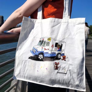 Puede incluir: Bolsa de tela blanca con una ilustración pintada a mano de un camión de helados con una persona dentro y un ratón sosteniendo un cono de helado. El camión es azul y blanco con las palabras "Let's Roll" y "Milk Shakes" en el lateral. El ratón es marrón y blanco. La bolsa tiene las palabras "JohnnyWeirdDraws" en la parte inferior.