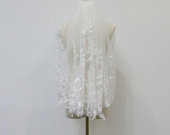 May include: A white bridal veil with delicate floral lace embroidery. The veil is displayed on a mannequin.