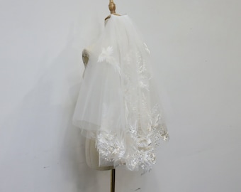 May include: A white bridal veil with delicate lace trim and a floral embellishment. The veil is displayed on a mannequin.