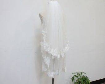 May include: A white, two-tiered bridal veil with delicate lace trim along the edges. The veil is displayed on a mannequin, showcasing its flowing, sheer fabric and intricate design. The lace adds a touch of elegance.