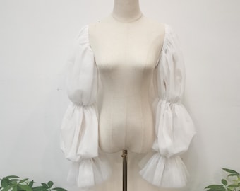 May include: White sheer long-sleeved blouse with puffed sleeves and ruffles. The sleeves are gathered at the elbow and wrist.