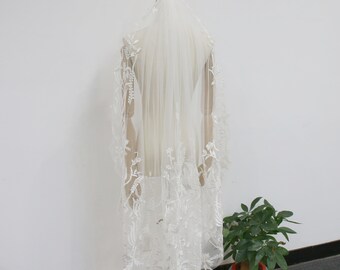 May include: A white bridal veil with delicate floral lace trim. The veil is displayed on a mannequin and has a soft, flowing texture.