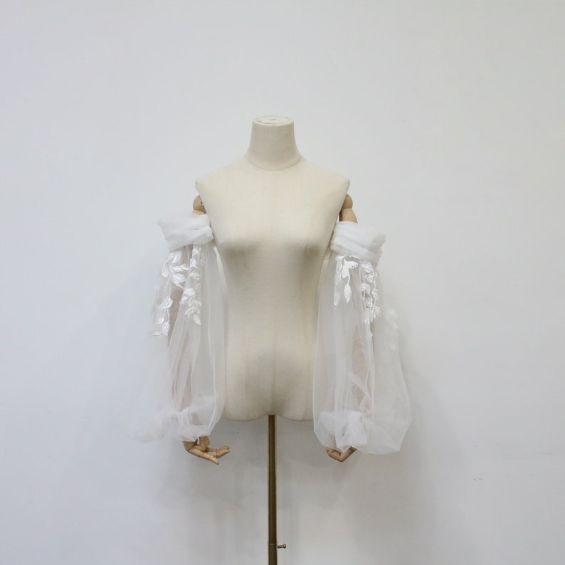 May include: White tulle off-the-shoulder sleeves with delicate floral lace embroidery. The sleeves are gathered at the wrist and have a puffed look.