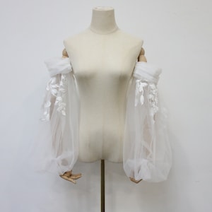 May include: White tulle off-the-shoulder sleeves with delicate floral lace embroidery. The sleeves are gathered at the elbow and have a flowing, romantic look.