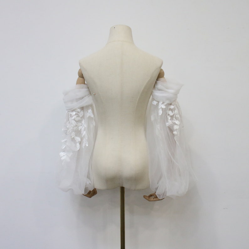 May include: White tulle bridal sleeves with delicate floral lace embroidery. The sleeves are gathered at the elbow and have a flowing, romantic look.
