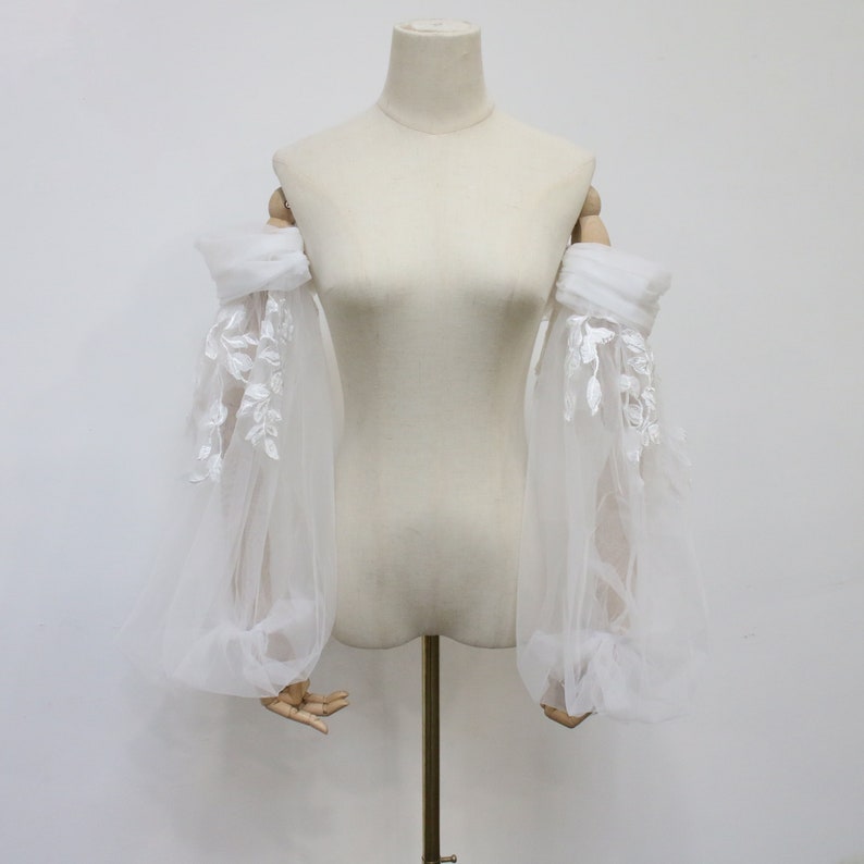 May include: A pair of white tulle sleeves with delicate floral lace embroidery. The sleeves are gathered at the elbow and have a flowing, romantic look.